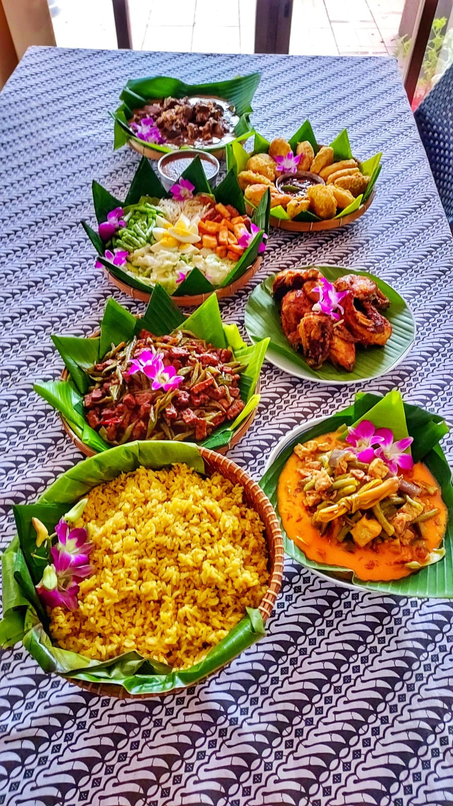 Tablecloth with black-and-white batik print carrying bowls of nasi kuning, rendang, sambal prawns and fried chicken in banana-leaf-lined baskets