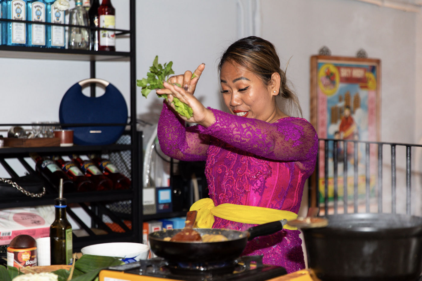 Chef Novy in a magenta lace kebaya tossing fresh herbs into a sizzling pan during a live private-cooking experience