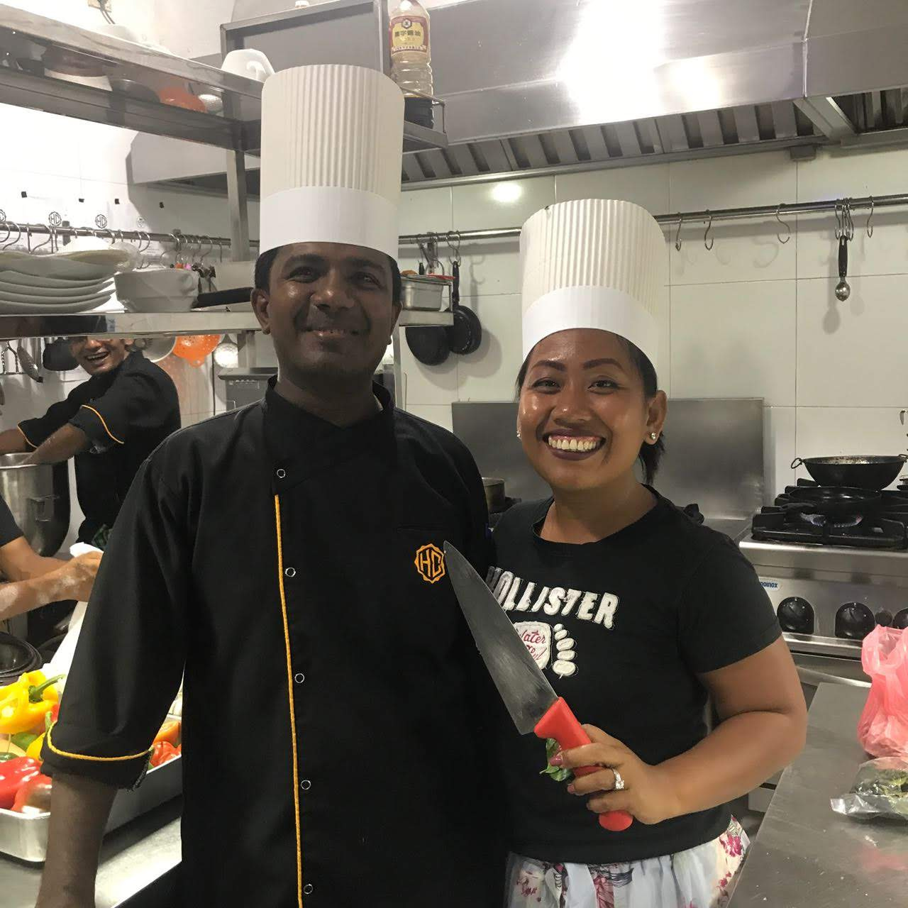 Guest Chef Novy beaming in a tall chef's hat beside the head chef in the professional kitchen at The Heritage Café & Bistro, Galle Fort, Sri Lanka
