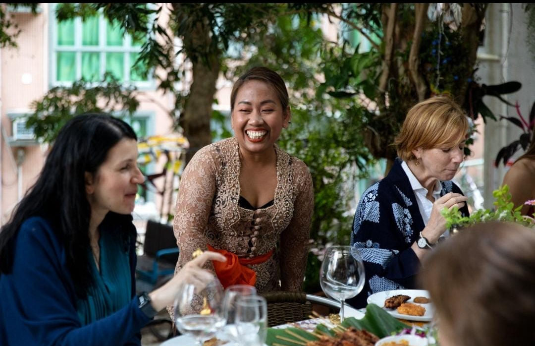 Chef Novy in a champagne-gold lace kebaya smiling as she serves guests around an outdoor table laid with satay and Indonesian dishes at a private event
